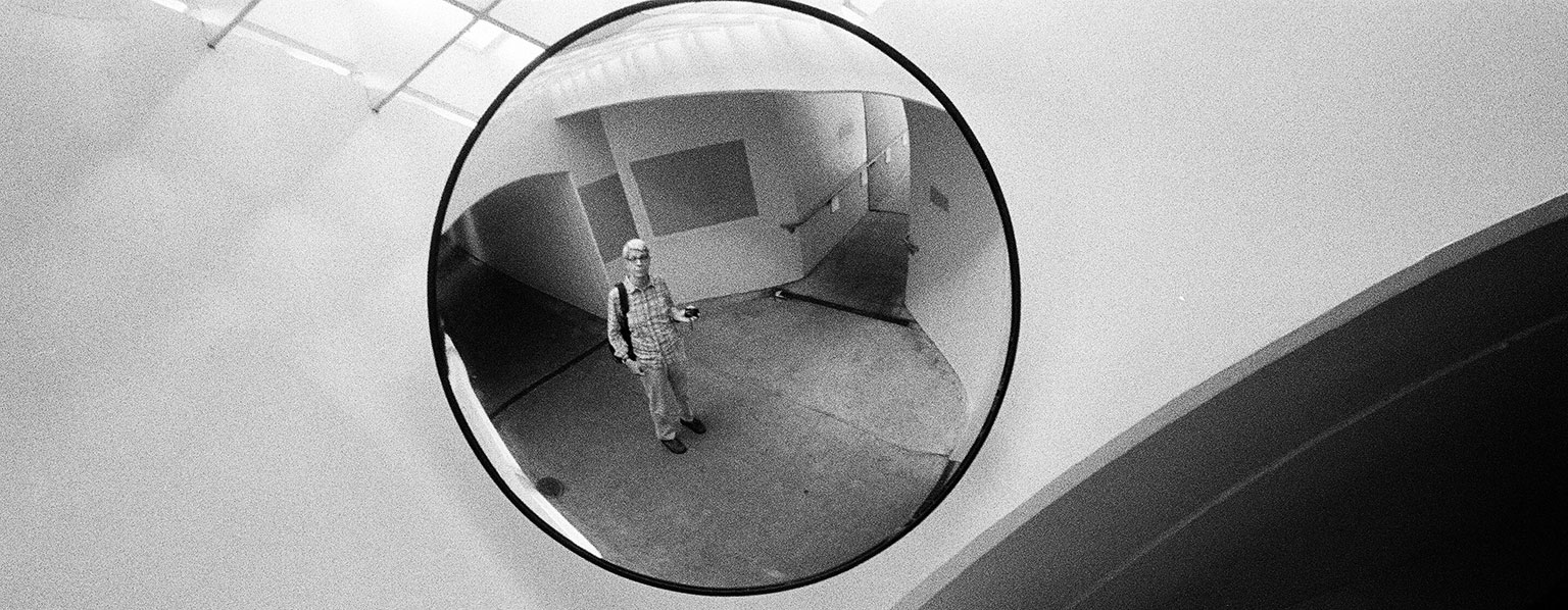 Black and white photo of a convex security mirror mounted on a wall, reflecting a person standing in an architectural space with curved walls and ramps. The individual wearing a backpack and holding a camera, appears centered in the mirror’s reflection, creating a surreal and distorted visual effect.